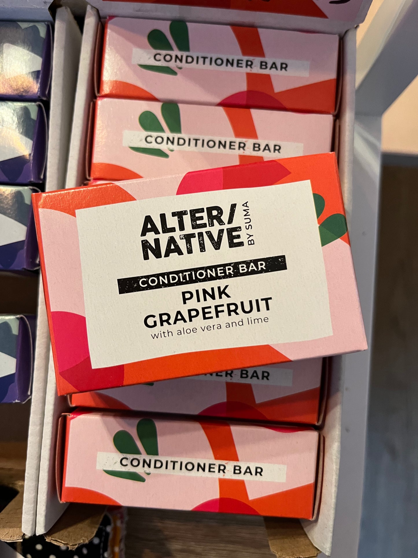 A rectangular brightly coloured pink, orange and green box containing a Pink Grapefruit, Aloe Vera & Lime scented conditioner bar sits on top of a row of more solid conditioner bars.