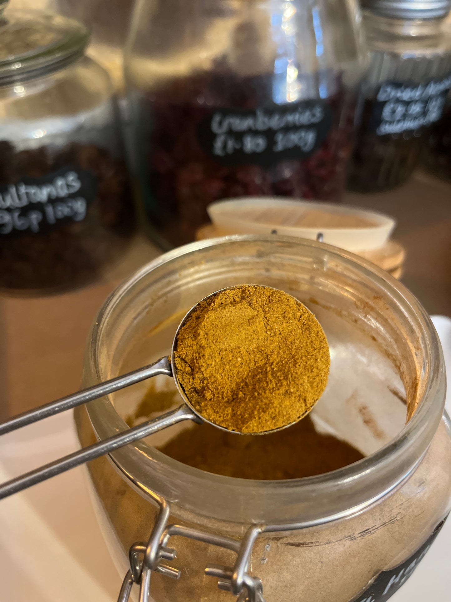 A scoop of warm golden brown spice is held over an open kilner jar in front of a row of jars filled with various dried fruit.