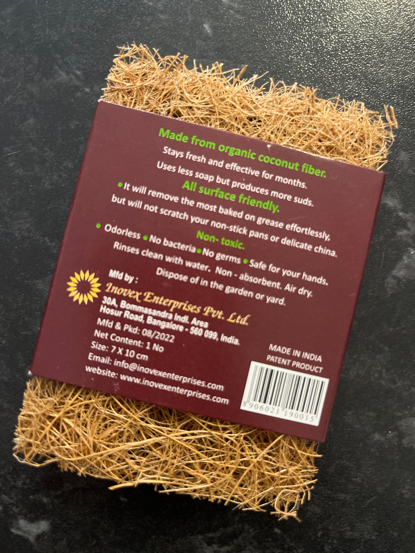 A coconut scrub pad or scourer. The reverse of a brown card sleeve with 'Made from organic coconut fibre. Stays fresh and effective for months. Uses less soap but produces more suds. All surface friendly. It will remove the most baked on grease effortlessly but will not scratch your non-stick pans or delicate china. Non-toxic. Odourless. No bacteria. No germs. Safe for your hands. Rinses clean with water. Non-absorbent. Air dry. Dispose of in the garden or yard.' printed in white and green.