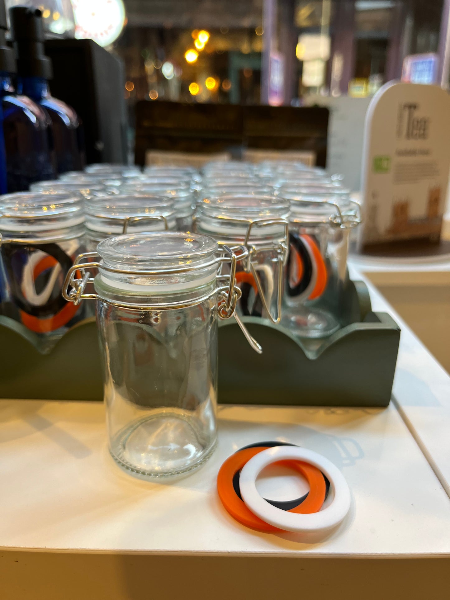 A round clear glass spice jar sits in front of a tray of similar jars, with spare lid seals in white, orange and black beside it.