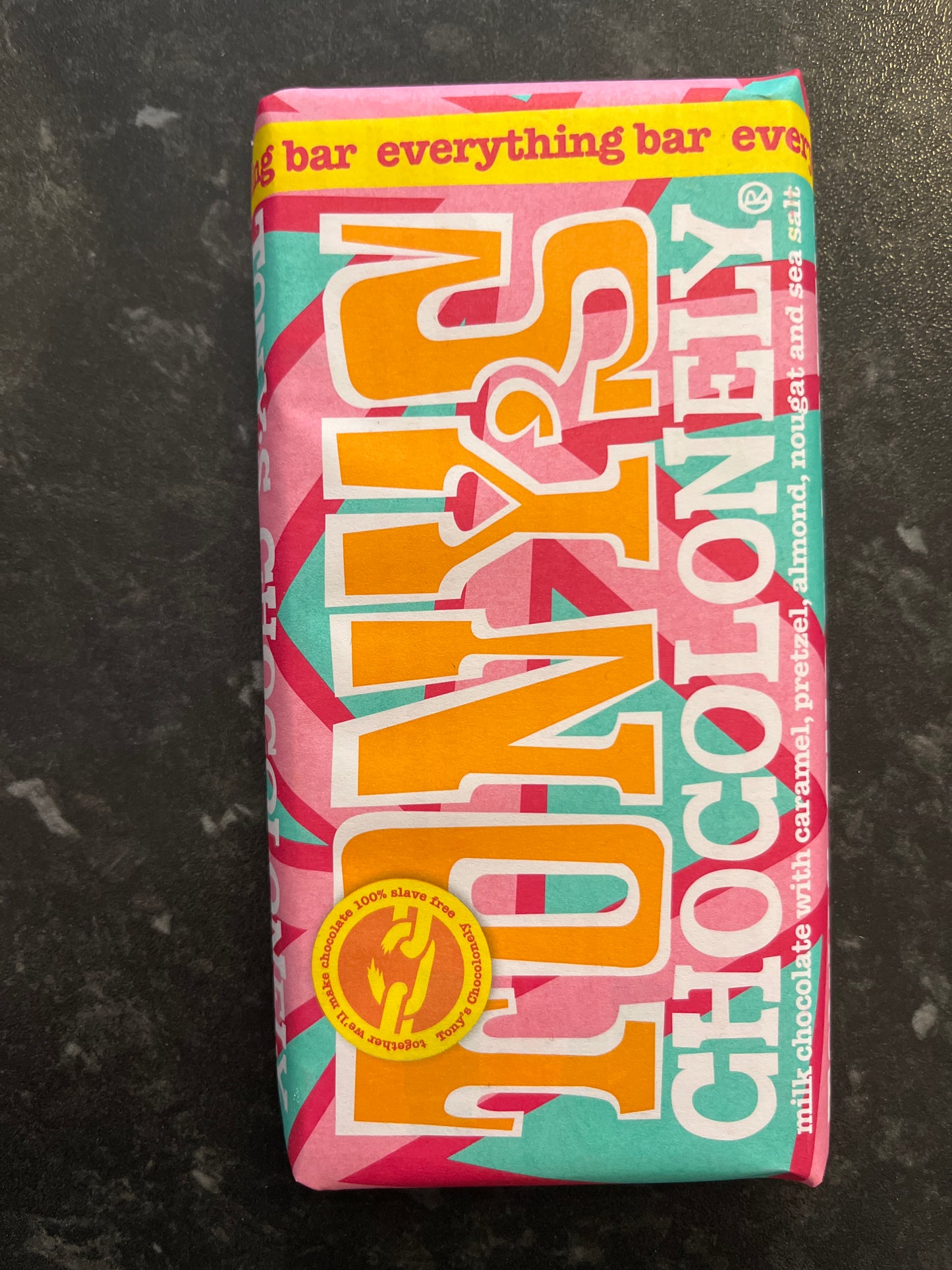 A large Everything Tony’s bar in a pink & aqua paper wrapper on a dark counter