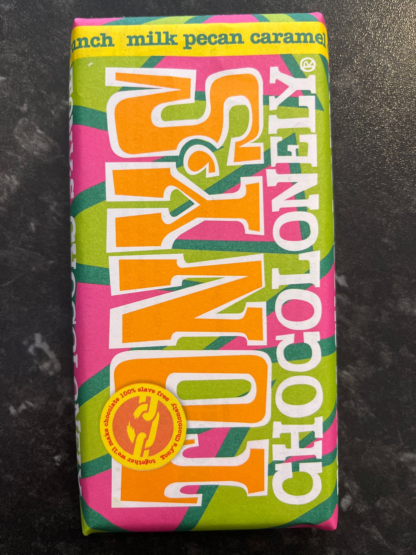 A Tony’s Chocolonely Caramel Pecan bar in a pink & green paper wrapper on a dark counter
