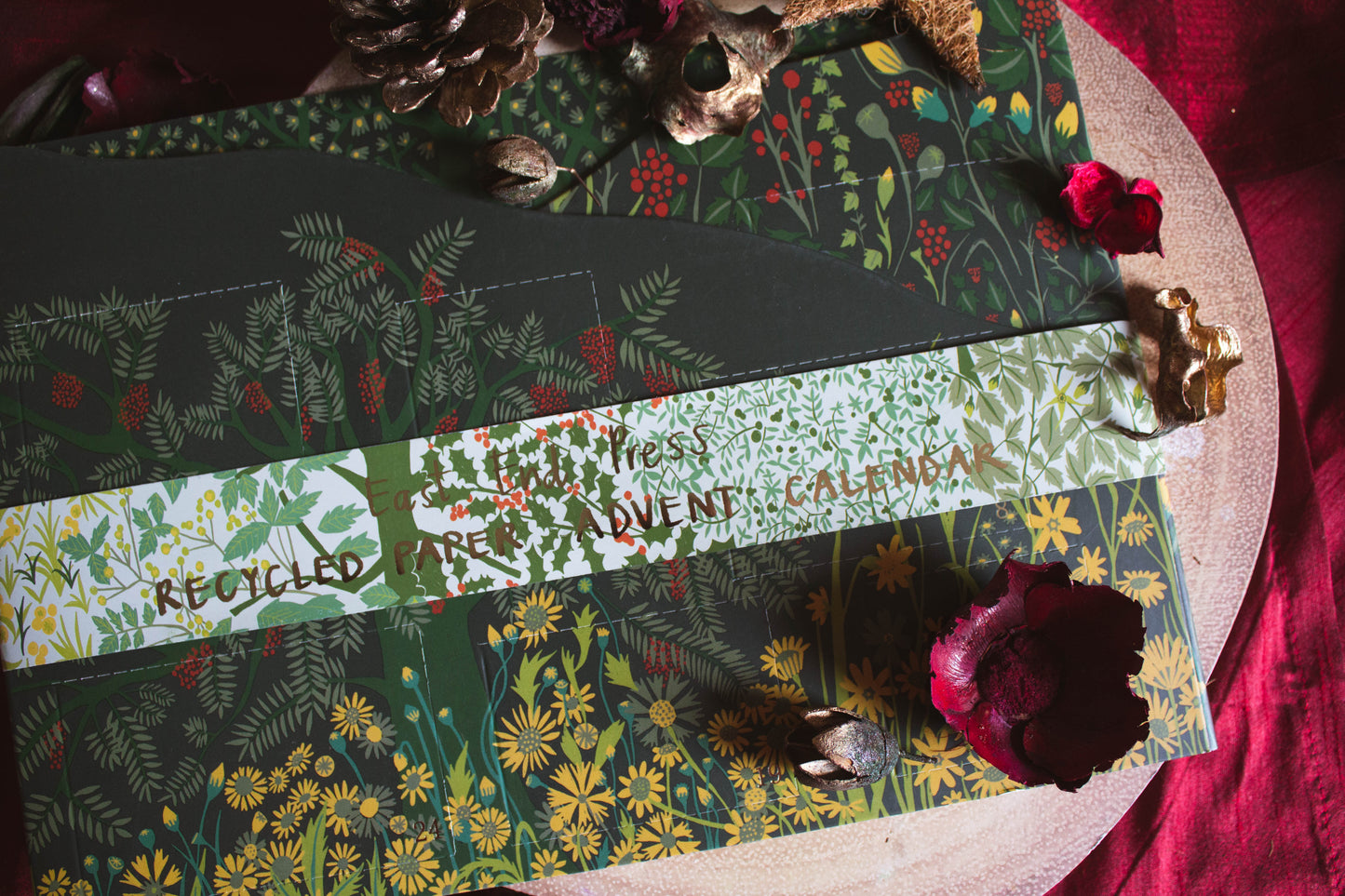 A deep green rectangular, fold-out advent calendar with a floral design is laying on a metallic circular tray on a red fabric background. There are red rose petals, pinecones, and gold leaves.