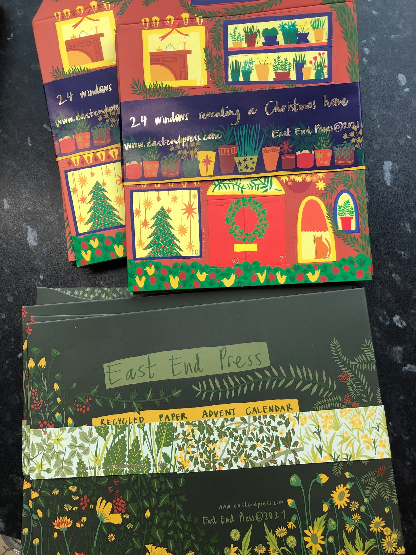 Two piles of advent calendars are laid on a counter, one above the other, the above is the house-shaped calendar and bellow is the green floral one.