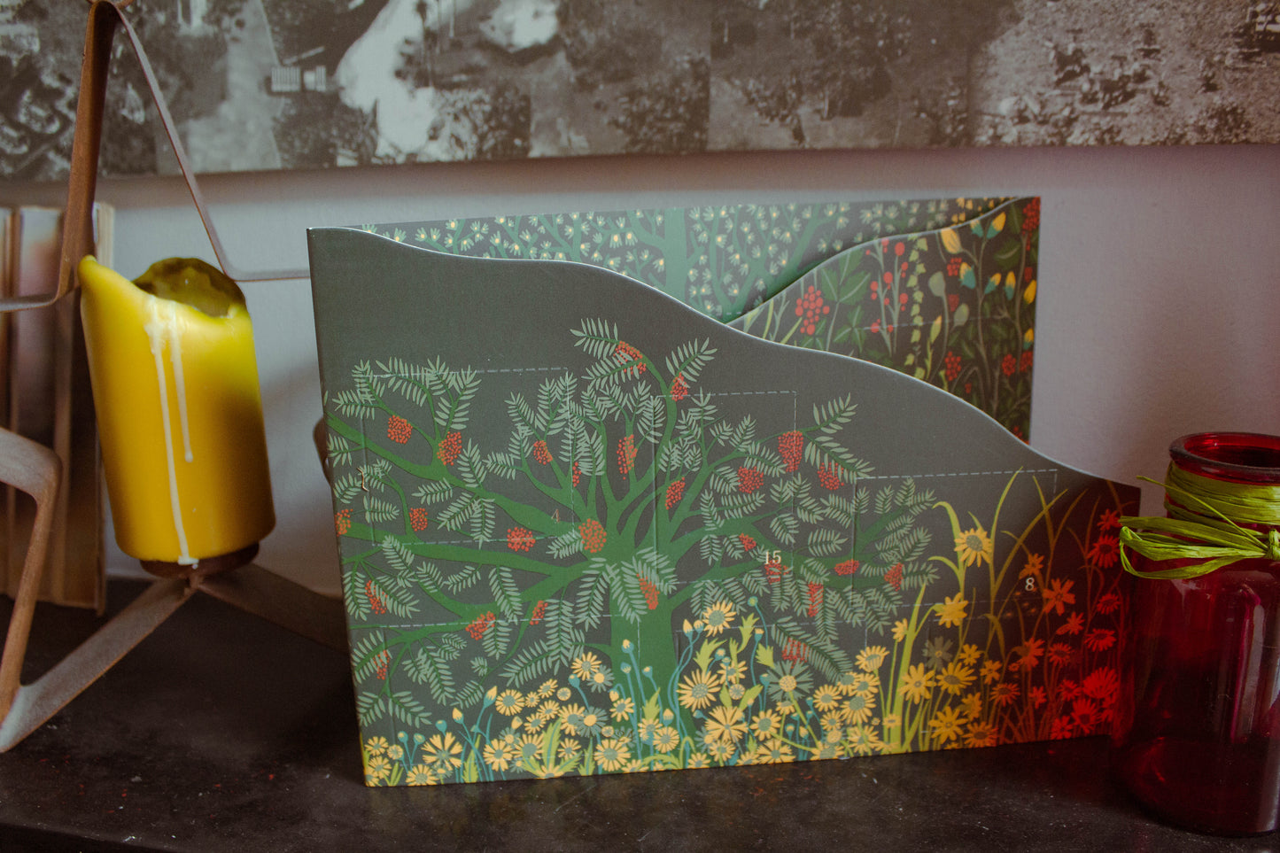 A picture of the Green floral advent calendar on a sideboard, a yellow candle the the left and a red jar to its right.