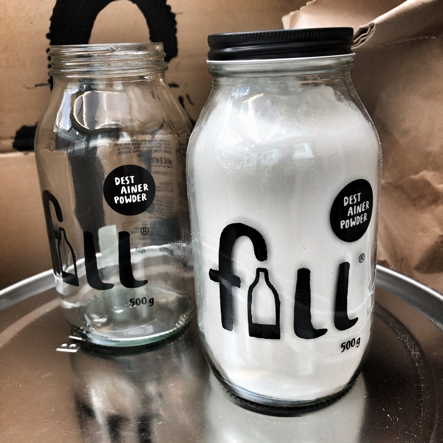 Two 500ml glass jars, one empty with the lid off and one filled with white eco cleaning powder with a black screw top lid on.