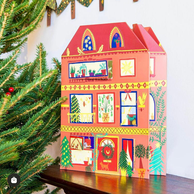 A house-shaped advent calendar is pictured, which has three stories and an antic with peachy orange walls, a deep peach/pink roof with varying sizes of windows and a door with as wreath on the bottom. Each window is highlighted with red, blue, or yellow and festivities can be seen within.