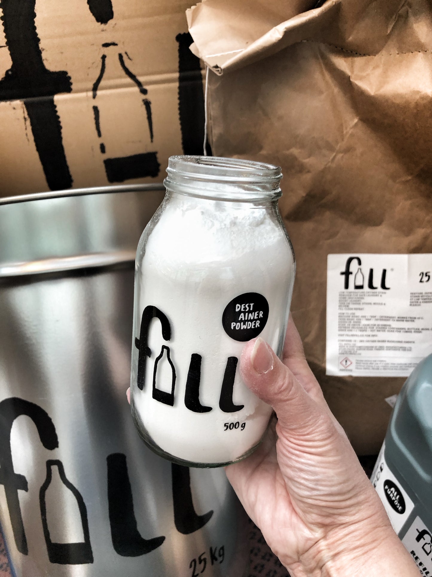 A hand holds an open jar of eco cleaning powder in front of a silver refill canister and brown paper sack of plastic free cleaning refills.