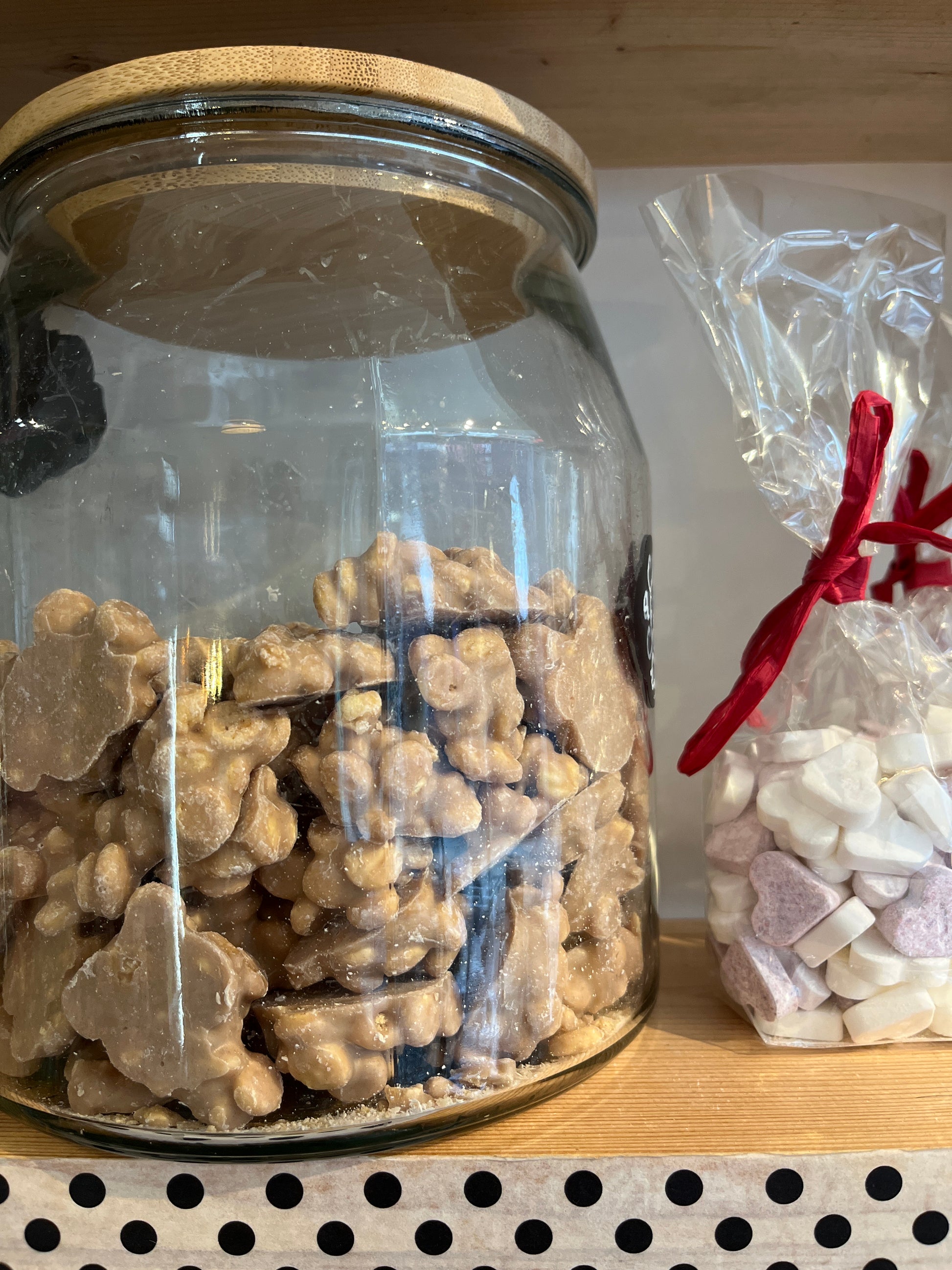 A big jar with a wooden top half filled with chocolate apple pie clusters sits on a shelf next to clear compostable bags of pale heart shaped sweets tied with red ribbon.