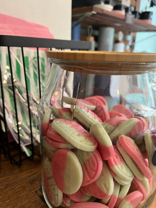 A large jar with a wooden lid containing oval pink and green vegan watermelon foam sweets. A container nearby holds pink and green paper bags and silver tongs.