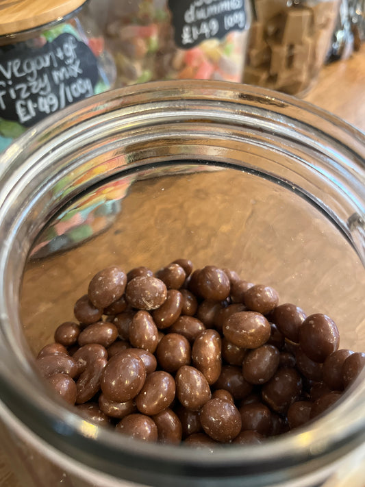 Chocolate coated peanuts are just seen in the open mouth of a glass jar with various coloured sweets in jars glimpsed behind.