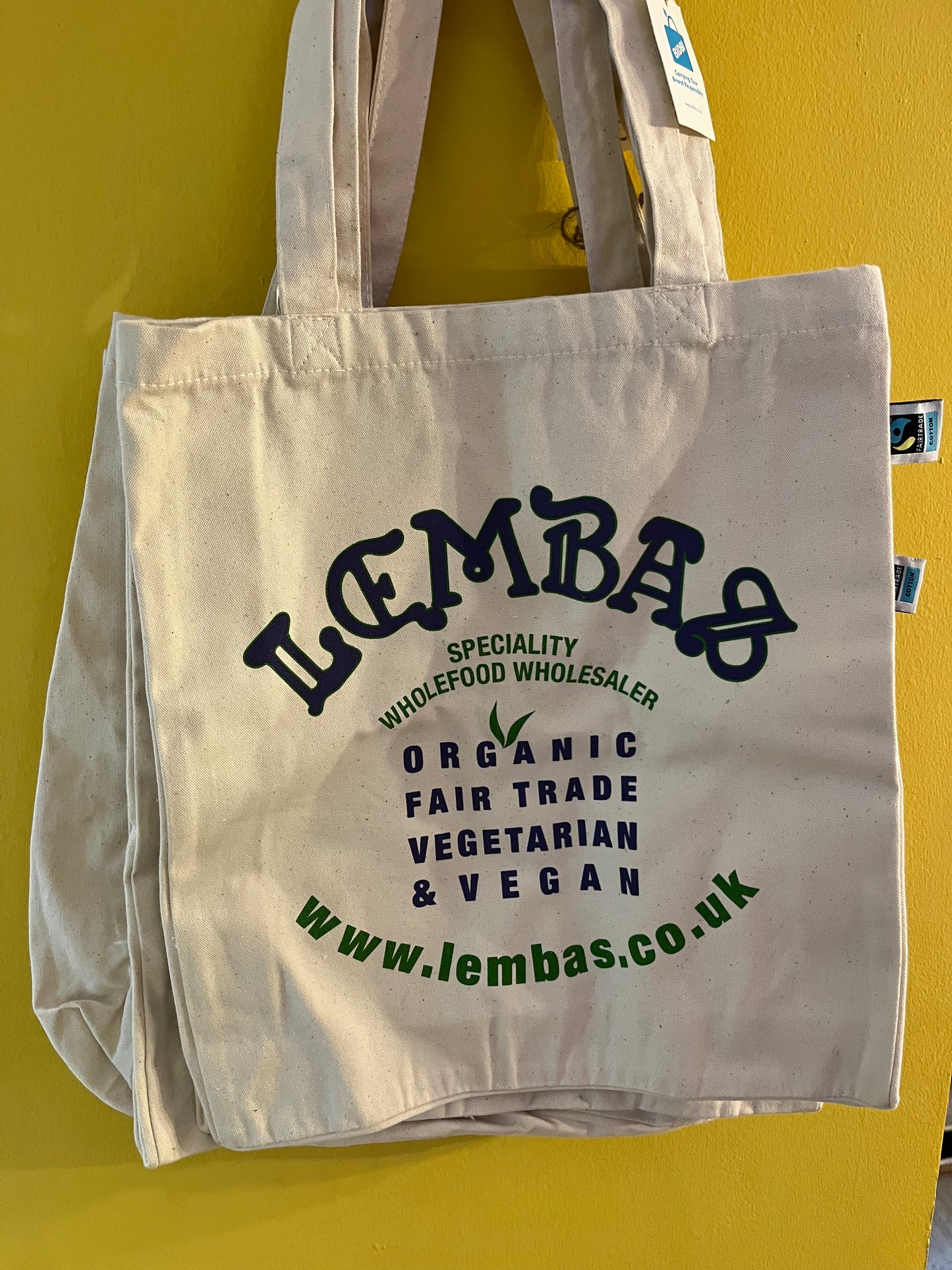 A natural cotton bag with wide shoulder straps, printed in dark blue and green to read ‘Lembas, Specialty Wholefood Wholesaler, Organic, Fairtrade, Vegetarian, & Vegan’