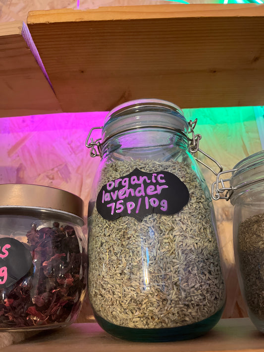 A jar filled with dried flowers labelled 'organic lavender 75p per 10g' in pink chalk pen on a black label sits on a wooden shelf with pink and green lighting coming down from above.