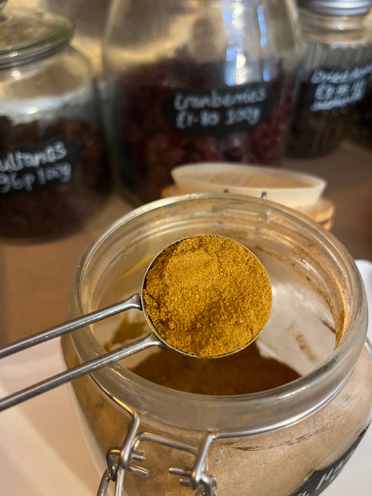 A scoop of warm golden brown spice is held over an open kilner jar in front of a row of jars filled with various dried fruit.